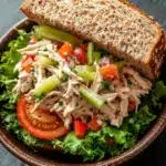 Chicken salad sandwich with veggies - the image shows a bowl of salad with a slice of bread on top. the salad is made up of lettuce, cherry tomatoes, celery, and other vegetables. the bread is toasted and appears to be freshly baked. the bowl is made of dark brown ceramic and is placed on a dark grey surface. the background is blurred, making the salad the focal point of the image.