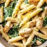 Chicken spinach skillet pasta with lemon parmesan - the image is a close-up of a bowl of pasta dish. the pasta appears to be penne or penne, and it is cooked al dente. the dish is garnished with fresh spinach leaves and chunks of chicken. the chicken is cooked to a golden brown color and is sprinkled with grated parmesan cheese. the bowl is made of ceramic and is sitting on a white marble countertop.
