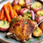 Chicken thigh dinner dutch oven - the image shows a plate of food with a serving of roasted chicken and vegetables. the chicken is golden brown and appears to be seasoned with herbs and spices. the vegetables on the plate include carrots, potatoes, and red onions. the plate is blue and has a textured surface. the background is white and the overall color scheme of the image is warm and inviting.