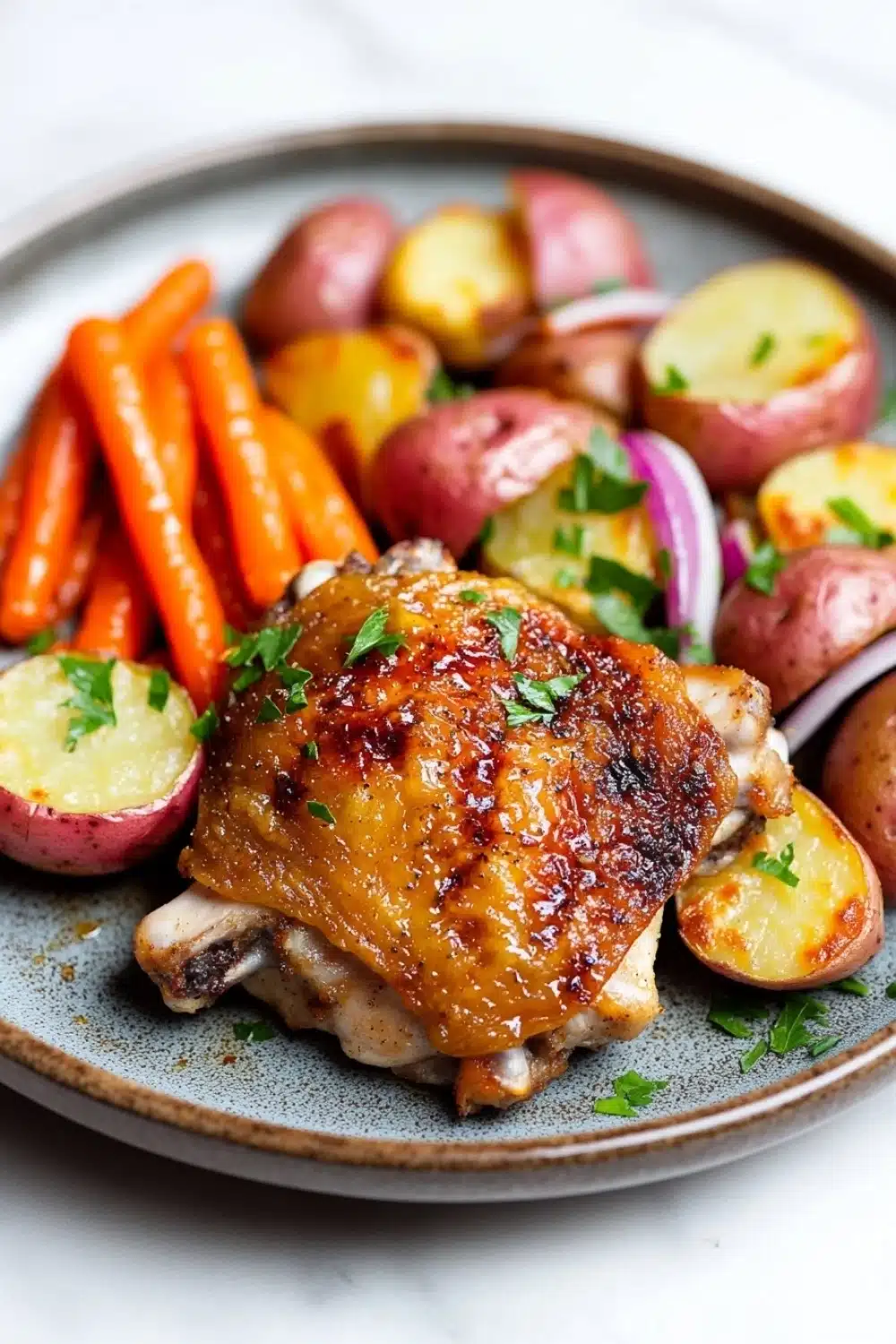 Chicken thigh dinner dutch oven - the image shows a plate of food with a serving of roasted chicken and vegetables. the chicken is golden brown and appears to be seasoned with herbs and spices. the vegetables on the plate include carrots, potatoes, and red onions. the plate is blue and has a textured surface. the background is white and the overall color scheme of the image is warm and inviting.