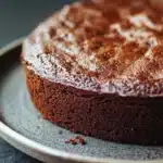 Chocolate banana yoghurt cake - the image is a close-up of a round chocolate cake on a grey plate. the cake appears to be freshly baked and has a golden brown crust on top. the top of the cake is covered in a generous layer of powdered sugar, giving it a shiny and shiny appearance. the plate is sitting on a dark grey surface, and there is a yellow bowl in the background. the overall mood of the image is warm and inviting.