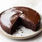 Chocolate cake japanese - the image shows a round chocolate cake on a white marble plate. the cake is covered in a thick layer of chocolate frosting and has a slice taken out of it. the frosting is smooth and glossy, and the cake appears to be freshly baked. the plate has a gold rim and the background is blurred, making the cake the focal point of the image.