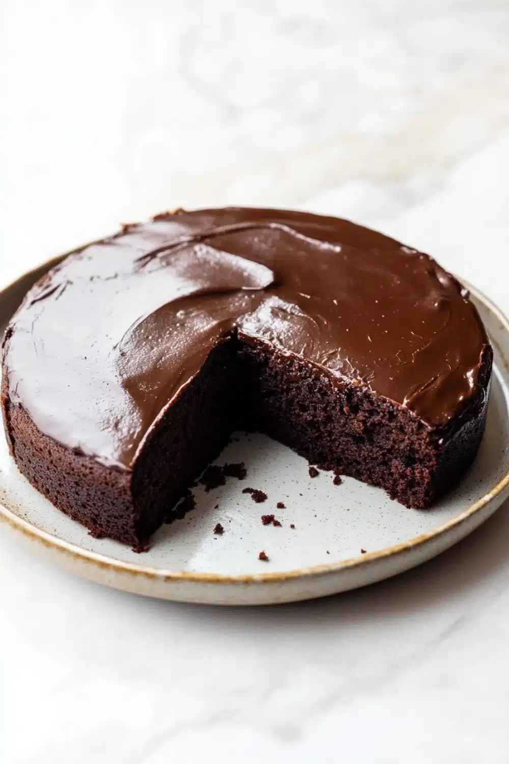 Chocolate cake japanese - the image shows a round chocolate cake on a white marble plate. the cake is covered in a thick layer of chocolate frosting and has a slice taken out of it. the frosting is smooth and glossy, and the cake appears to be freshly baked. the plate has a gold rim and the background is blurred, making the cake the focal point of the image.
