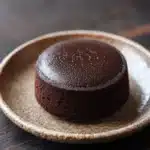 Chocolate cake japanese - the image shows a round chocolate cake on a beige plate. the cake appears to be moist and has a smooth texture. it is covered in a light dusting of powdered sugar, giving it a shiny appearance. the plate is placed on a dark wooden surface, and the background is blurred, making the cake the focal point of the image.