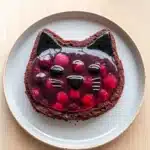 Chocolate cake jelly cat - the image is of a round chocolate cake with a cat face on top. the cake is on a white plate with a light blue rim. the cat face is made up of black cat ears and is covered in a layer of red cherries. the cherries are arranged in a circular pattern around the cat's face. the background is a light beige color.