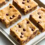 Chocolate chip cookie dough bars - the image shows a tray of freshly baked chocolate chip cookie bars. the bars are golden brown and have a crumbly texture. they are arranged in a single layer on the tray, with some overlapping each other. the chocolate chips are scattered across the top of the bars, adding a pop of color and texture. the tray is lined with parchment paper, and the background is a light blue countertop.
