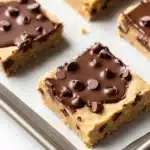 Chocolate chip cookie dough bars - the image shows a baking tray with six square-shaped bars on it. the bars are golden brown in color and have a crumbly texture. they are covered in a thick layer of chocolate chips, which are scattered across the top of each bar. the chocolate chips are dark brown and appear to be freshly baked. the tray is lined with parchment paper and there are a few crumbs scattered around the tray. the background is white.