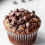 Chocolate kodiak muffin - the image is of a freshly baked chocolate muffin on a white marble countertop. the muffin is round and has a golden brown color. it is topped with chocolate chips and nuts, giving it a crunchy texture. there are a few scattered chocolate chips on top of the muffin, adding a pop of color to the image. the background is blurred, but it appears to be a kitchen countertop with a window on the left side.