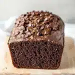 Chocolate zucchini yogurt bread - the image shows a freshly baked loaf of bread on a wooden cutting board. the bread appears to be dark brown in color and has a slightly crumbly texture. it is topped with chocolate chips, giving it a shiny appearance. the cutting board is placed on a white marble countertop. the background is blurred, making the bread the focal point of the image.
