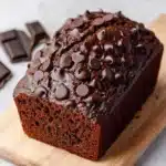 Chocolate zucchini yogurt bread - the image shows a rectangular loaf of chocolate cake on a wooden cutting board. the cake appears to be freshly baked and has a rich, dark brown color. the top of the cake is covered in a thick layer of chocolate shavings, which are scattered across the surface. there are also a few pieces of chocolate scattered around the cutting board, suggesting that the cake has been freshly baked. the cutting board is placed on a white marble countertop.