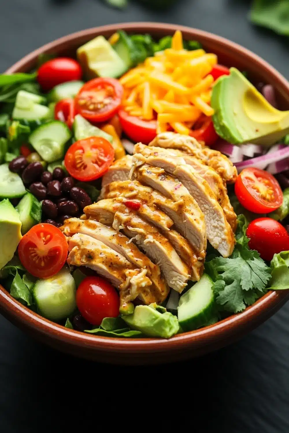 Chopped chicken salad with creamy chipotle dressing - the image is a close-up of a large bowl of salad. the salad is made up of various ingredients such as sliced chicken, sliced avocado, cherry tomatoes, black beans, red onions, and cilantro. the chicken is cooked to a golden brown color and appears to be seasoned with herbs and spices. the avocado is cut into thin slices and is placed on top of the salad, while the cherry tomatoes are scattered throughout. the black beans are black and appear to be kidney beans. the red onions are cut into small cubes and are arranged in a circular pattern around the chicken. the cilantro is scattered throughout the salad and adds a pop of green color to the dish. the bowl is sitting on a dark table with a blurred background.