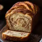 Cinnamon bread - the image shows a freshly baked loaf of bread on a wooden cutting board. the bread has a golden brown crust and a swirl pattern on the top. a slice of the bread is cut in half, revealing the soft, fluffy interior. the cutting board is placed on a dark wooden table with a white cloth napkin on the right side. the background is blurred, but it appears to be a kitchen countertop.