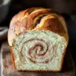 Cinnamon bread - the image shows a freshly baked loaf of bread on a wooden cutting board. the bread is golden brown in color and has a swirl pattern on top. the swirl appears to be made with cinnamon or sugar, giving it a warm and inviting appearance. the cutting board is placed on a rustic wooden table, with a few sprigs of cinnamon scattered around it. the background is blurred, making the bread the focal point of the image.