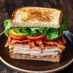 Club sandwich - the image is of a sandwich on a brown plate. the sandwich is made with two slices of toasted bread, filled with sliced turkey, bacon, lettuce, tomato, and pickles. the turkey is cooked to a golden brown color and appears to be seasoned with herbs and spices. the lettuce is a mix of green and red, and the pickles are a bright yellow. the plate is sitting on a wooden table with a rustic background.