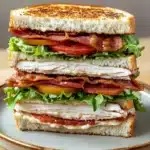 Club sandwich - the image is of a sandwich on a plate. the sandwich is made with two slices of toasted bread, filled with a variety of ingredients. on top of the bread, there are slices of bacon, lettuce, tomato, and chicken. the bacon is crispy and golden brown, and the lettuce is a vibrant green. the chicken is white and appears to be cooked to perfection. the plate is a light brown color and is placed on a wooden table.
