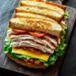 Club sandwich on french bread - the image shows a sandwich cut in half on a wooden cutting board. the sandwich is made with two slices of toasted bread, filled with sliced turkey, lettuce, tomato, and cheese. the bread is golden brown and appears to be crispy on the outside. the cheese is melted and oozing out of the sides of the sandwich. the cutting board is placed on a dark grey textured surface, and there is a small wooden spoon next to it.