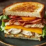 Club sandwich with chicken - the image is of a sandwich on a green plate. the sandwich is made with two slices of toasted bread, filled with grilled cheese, bacon, and lettuce. the cheese is melted and oozing out of the sides. the bacon is crispy and golden brown, and the lettuce is a vibrant green color. the plate is sitting on a wooden table with a blurred background.