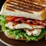 Club sandwich with chicken - the image shows a sandwich on a brown plate. the sandwich is made with two slices of toasted bread, filled with grilled chicken, bacon, and lettuce. the chicken is cooked to a golden brown color and appears to be seasoned with herbs and spices. the bacon is crispy and golden brown, and the lettuce is a vibrant green. there is a small bowl of cherry tomatoes in the background. the plate is sitting on a wooden table.