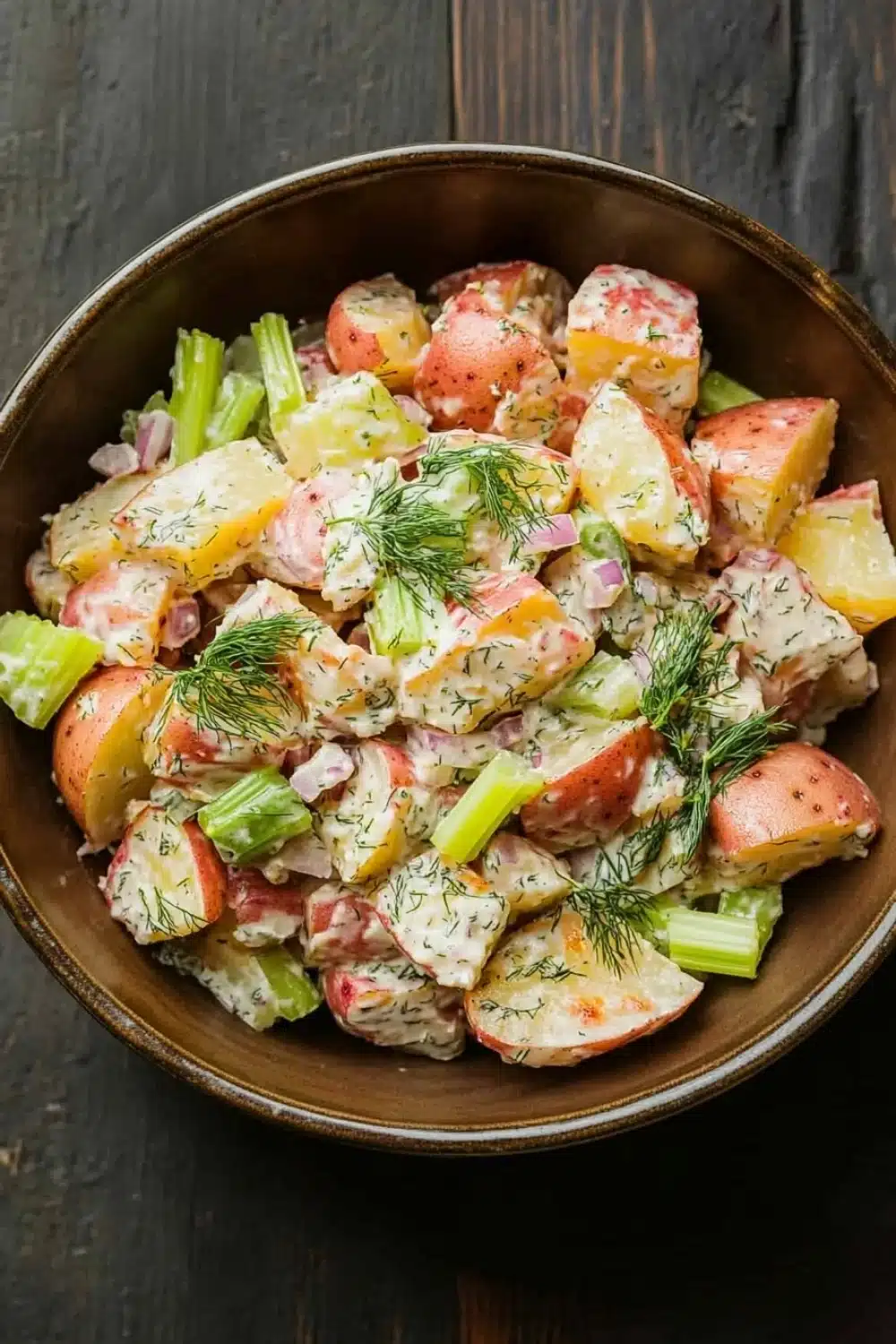 Creamy dill red potato salad - the image is a close-up of a bowl of potato salad. the bowl is made of dark brown ceramic and is placed on a wooden table. the salad is made up of diced potatoes, celery, and other vegetables, all mixed together in a creamy dressing. the potatoes are cut into small cubes and are arranged in a circular pattern. the dressing is drizzled over the top of the potatoes and is garnished with a sprig of dill. the overall appearance of the salad is fresh and appetizing.