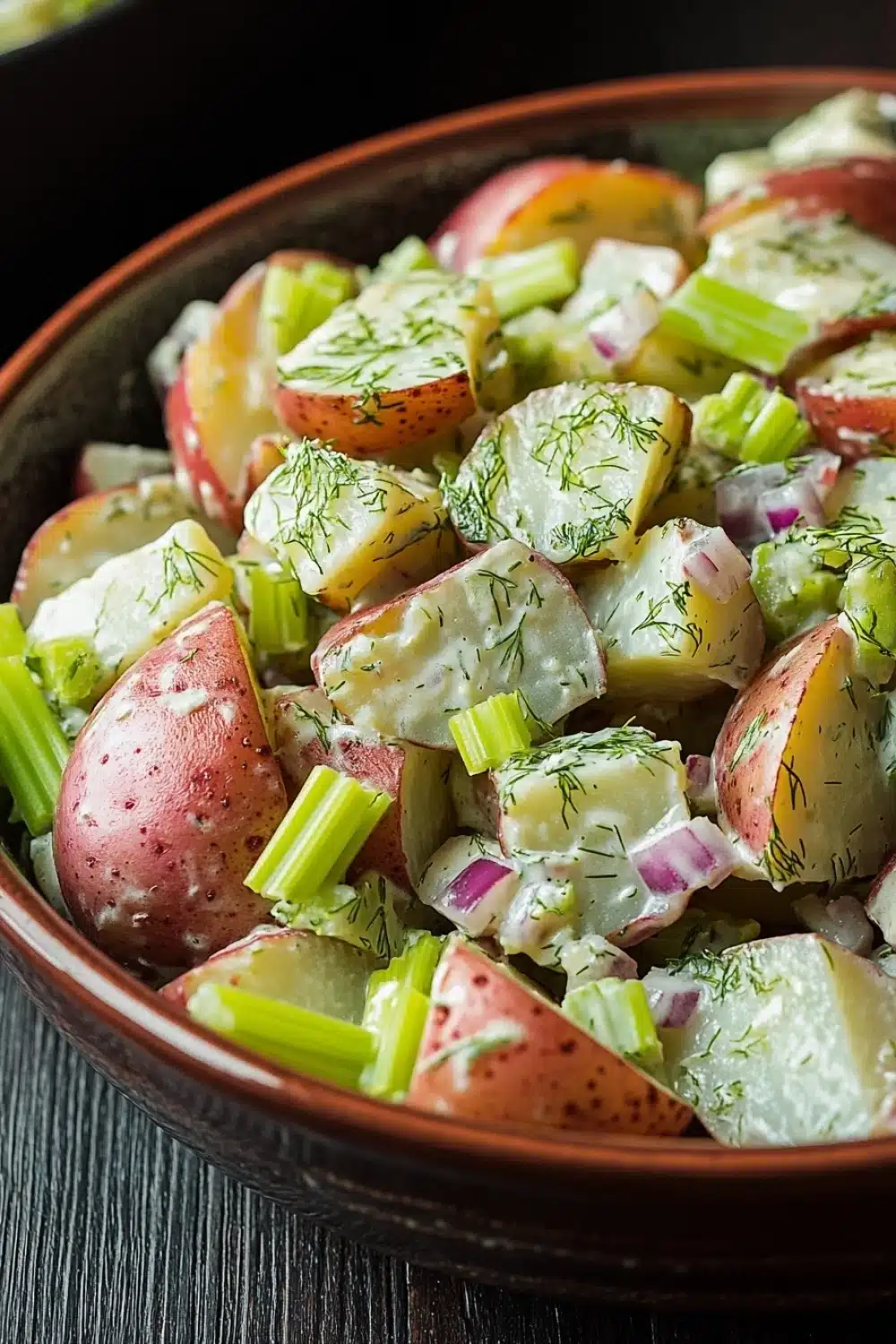Creamy dill red potato salad - the image is a close-up of a bowl of salad. the salad is made up of diced potatoes, red onions, and green celery. the potatoes are cut into small cubes and are arranged in a circular pattern. the celery is scattered throughout the salad, adding a pop of green color to the dish. the bowl is made of dark brown ceramic and is sitting on a wooden table. the background is blurred, but it appears to be a kitchen countertop.