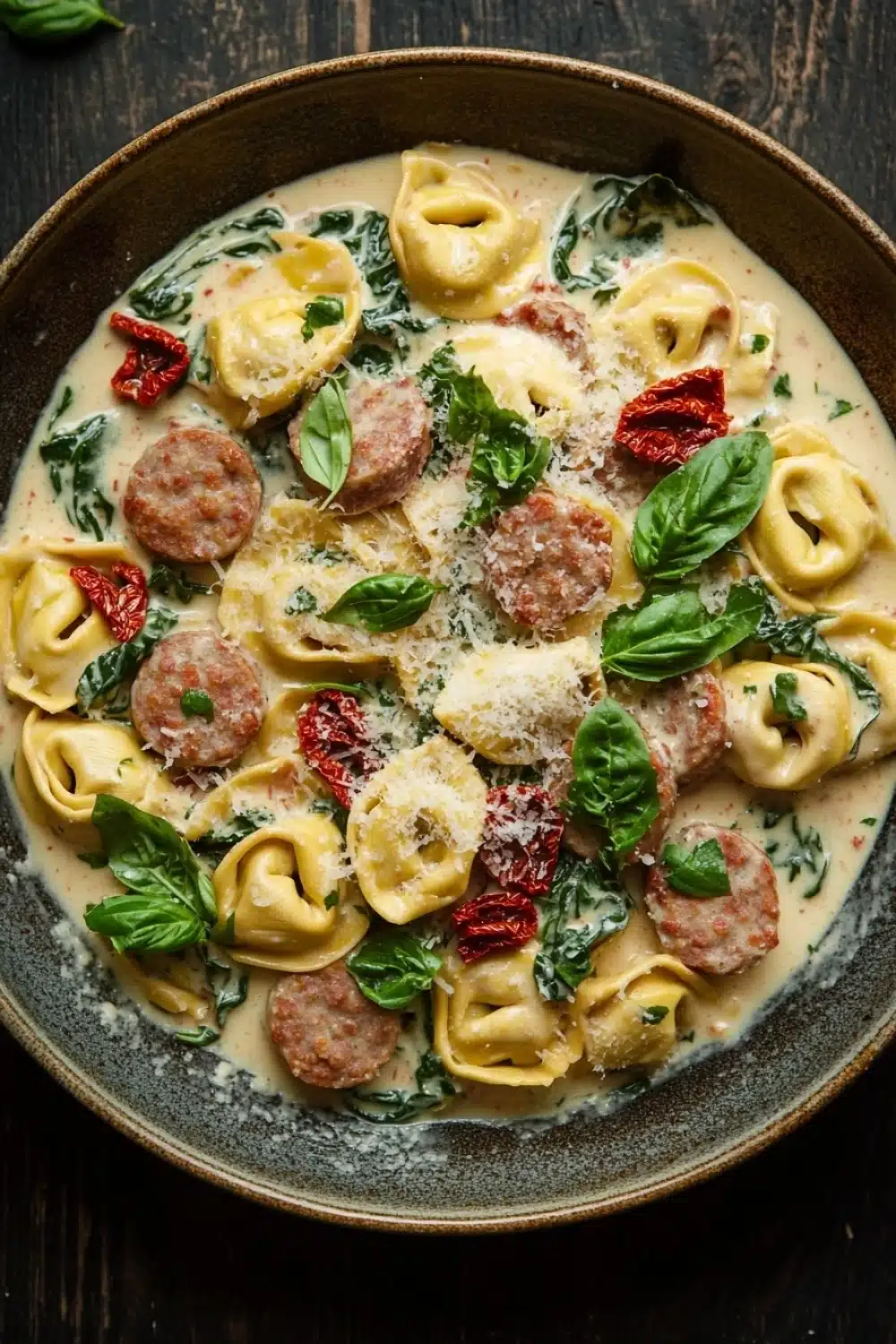 Creamy italian sausage tortellini skillet - the image is a close-up of a bowl of pasta dish. the dish appears to be a type of tortellini, with a creamy white sauce and chunks of sausage scattered throughout. the sauce is garnished with fresh basil leaves and red chili peppers. the bowl is made of dark brown ceramic and is sitting on a wooden table. the overall color scheme of the dish is warm and inviting.