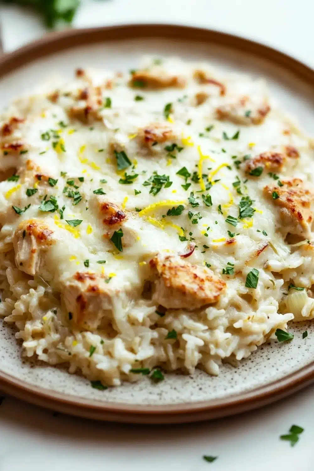 Creamy lemon chicken rice casserole - the image is a close-up of a plate of food. the plate is round and has a light brown rim. on top of the plate, there is a creamy white sauce with chunks of chicken and herbs scattered throughout. the sauce appears to be a creamy yellow color and is drizzled over the top. the chicken is cooked to a golden brown color and looks crispy on the outside. the dish is garnished with chopped parsley and a sprig of parsley. the background is blurred, but it seems to be an indoor setting with a white tablecloth.