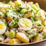 Creamy potato and egg salad - the image is a close-up of a bowl of potato salad. the bowl is made of brown ceramic and is placed on a white tablecloth. the salad is made up of diced potatoes, diced celery, red onions, and a dollop of hard-boiled eggs. the eggs are cut in half and are arranged in a circular pattern. there are also some chopped herbs scattered throughout the salad, adding a pop of green color. the overall appearance of the salad is fresh and appetizing.