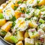 Creamy potato and egg salad - the image is a close-up of a bowl of potato salad. the salad is made up of diced potatoes, diced ham, diced celery, and hard-boiled eggs. the potatoes are yellow and appear to be seasoned with herbs and spices. the ham is cut into small cubes and is scattered throughout the salad. there are also some chopped parsley scattered throughout, adding a pop of green color to the dish. the bowl is made of dark brown ceramic and is sitting on a white marble countertop.