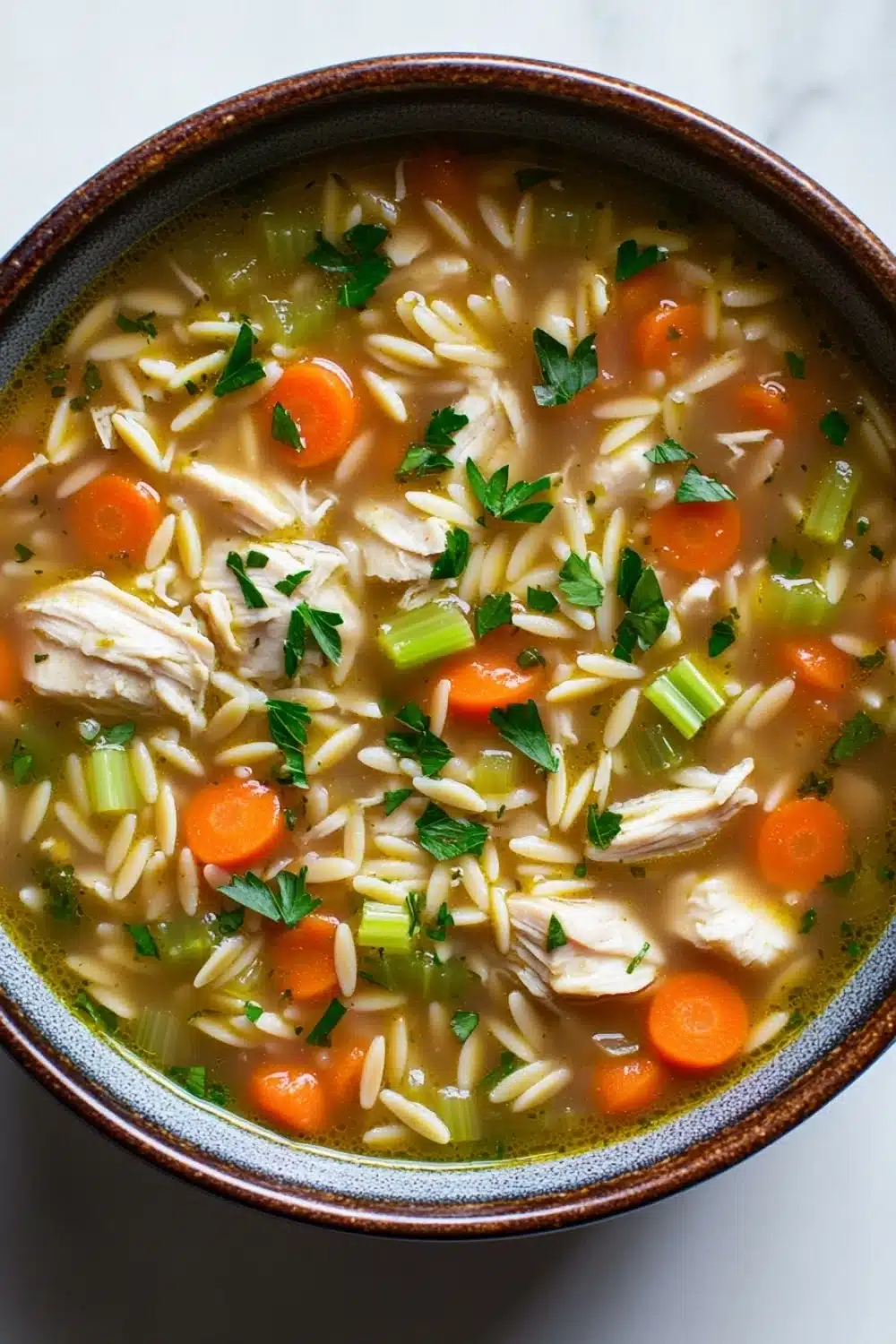 Crock pot chicken orzo soup - the image is a close-up of a bowl of soup. the soup appears to be a type of noodle soup with chunks of chicken, carrots, celery, and other vegetables mixed in. the bowl is made of ceramic and has a brown rim. the broth is a light brown color and looks thick and creamy. there are also a few sprigs of parsley scattered throughout the soup, adding a pop of green color to the dish. the background is a white marble countertop.