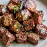 Crockpot garlic butter steak bites - the image shows a plate of cooked meat. the meat appears to be medium-rare and has a reddish-brown color. it is garnished with chopped herbs, likely thyme or rosemary. the plate is white with a speckled pattern, and the meat is arranged in a circular pattern around the edges. the background is blurred, but it seems to be a wooden table.