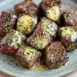 Crockpot garlic butter steak bites - the image shows a plate of cooked beef tenderloin cubes. the cubes are arranged in a circular pattern on a light blue plate with a speckled texture. each cube is covered in a creamy sauce and garnished with chopped herbs and spices. the meat appears to be cooked medium-rare and has a pinkish-red color. the plate is sitting on a wooden table with a white tablecloth in the background.