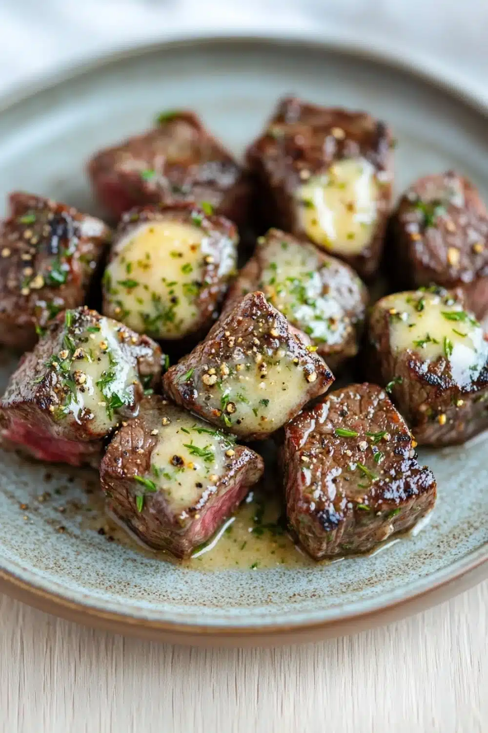 Crockpot garlic butter steak bites - the image shows a plate of cooked beef tenderloin cubes. the cubes are arranged in a circular pattern on a light blue plate with a speckled texture. each cube is covered in a creamy sauce and garnished with chopped herbs and spices. the meat appears to be cooked medium-rare and has a pinkish-red color. the plate is sitting on a wooden table with a white tablecloth in the background.