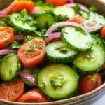 Cucumber tomato onion salad lemon - the image is a close-up of a bowl of salad. the salad is made up of sliced cucumbers, red onions, and cherry tomatoes. the cucumbers are cut into thin slices and are arranged in a circular pattern. the tomatoes are bright red and appear to be ripe and juicy. the red onions are sliced into thin strips and are scattered throughout the salad, adding a pop of color to the dish. there are also some chopped herbs and cilantro scattered throughout. the bowl is made of dark brown ceramic and is sitting on a white marble countertop.