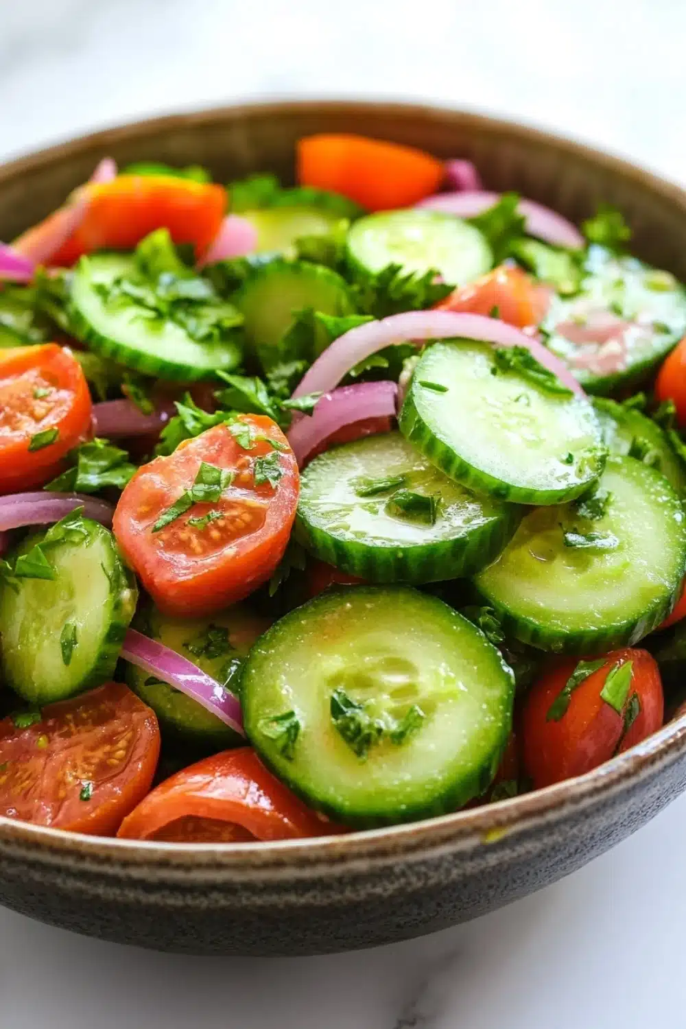 Cucumber tomato onion salad lemon - the image is a close-up of a bowl of salad. the salad is made up of sliced cucumbers, red onions, and cherry tomatoes. the cucumbers are cut into thin slices and are arranged in a circular pattern. the tomatoes are bright red and appear to be ripe and juicy. the red onions are sliced into thin strips and are scattered throughout the salad, adding a pop of color to the dish. there are also some chopped herbs and cilantro scattered throughout. the bowl is made of dark brown ceramic and is sitting on a white marble countertop.