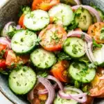 Cucumber tomato onion salad lemon - the image is a close-up of a bowl of salad. the salad is made up of sliced cucumbers, red onions, and cherry tomatoes. the cucumbers are cut into thin slices and are arranged in a circular pattern. the tomatoes are bright red and appear to be seasoned with herbs and spices. the red onions are sliced into thin strips and are scattered throughout the salad, adding a pop of color to the dish. the bowl is made of dark brown ceramic and is sitting on a white marble countertop.