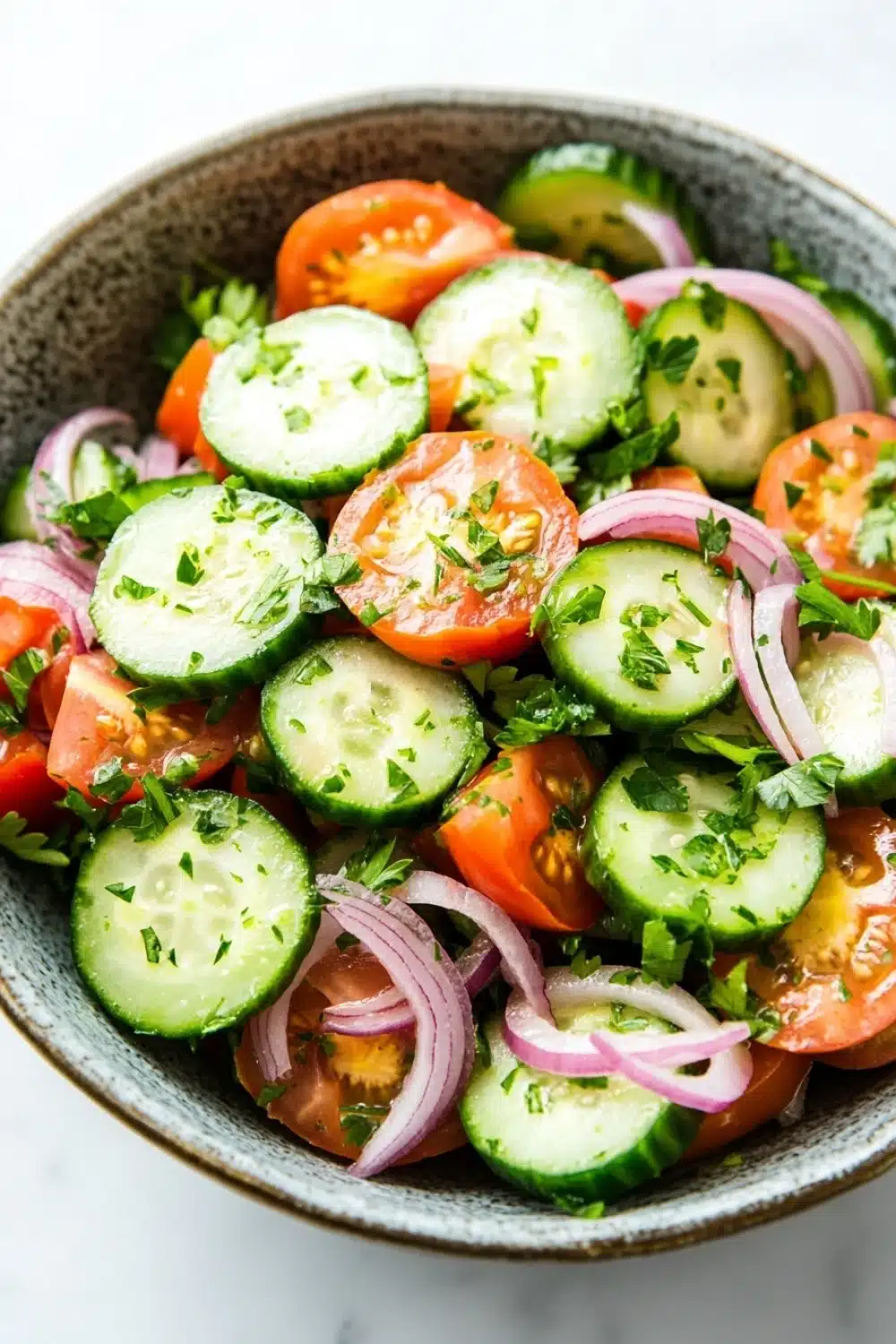 Cucumber tomato onion salad lemon - the image is a close-up of a bowl of salad. the salad is made up of sliced cucumbers, red onions, and cherry tomatoes. the cucumbers are cut into thin slices and are arranged in a circular pattern. the tomatoes are bright red and appear to be seasoned with herbs and spices. the red onions are sliced into thin strips and are scattered throughout the salad, adding a pop of color to the dish. the bowl is made of dark brown ceramic and is sitting on a white marble countertop.