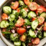Cucumber tomato salad lebanese - the image is a close-up of a bowl of salad. the bowl is made of ceramic and is placed on a white marble countertop. the salad is made up of sliced cucumbers, cherry tomatoes, red onions, and fresh herbs. the tomatoes are cut into small cubes and are arranged in a circular pattern. the herbs are scattered throughout the salad, adding a pop of green color to the dish. there are also a few sprigs of mint scattered around the bowl. the overall appearance of the salad is fresh and vibrant.