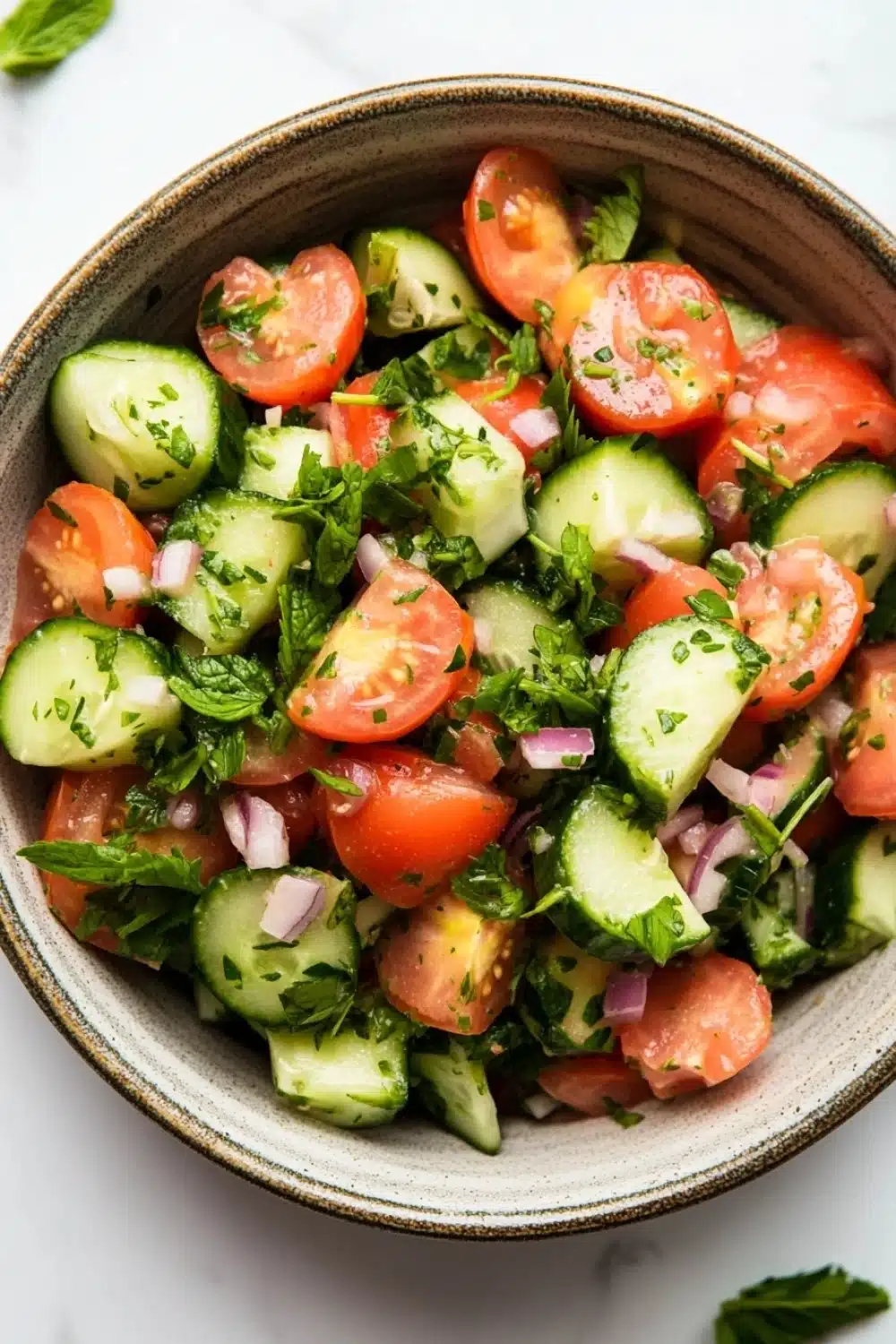 Cucumber tomato salad lebanese - the image is a close-up of a bowl of salad. the bowl is made of ceramic and is placed on a white marble countertop. the salad is made up of sliced cucumbers, cherry tomatoes, red onions, and fresh herbs. the tomatoes are cut into small cubes and are arranged in a circular pattern. the herbs are scattered throughout the salad, adding a pop of green color to the dish. there are also a few sprigs of mint scattered around the bowl. the overall appearance of the salad is fresh and vibrant.