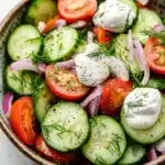 Cucumber tomato salad vinegar sour cream - the image is a close-up of a colorful salad in a bowl. the salad is made up of sliced cucumbers, red onions, cherry tomatoes, and dill leaves. the cucumbers are arranged in a circular pattern, with some overlapping each other. the tomatoes are bright red and appear to be ripe, while the onions are sliced into thin strips. the dill is scattered throughout the salad, adding a pop of green color to the dish. the bowl is sitting on a white marble countertop.