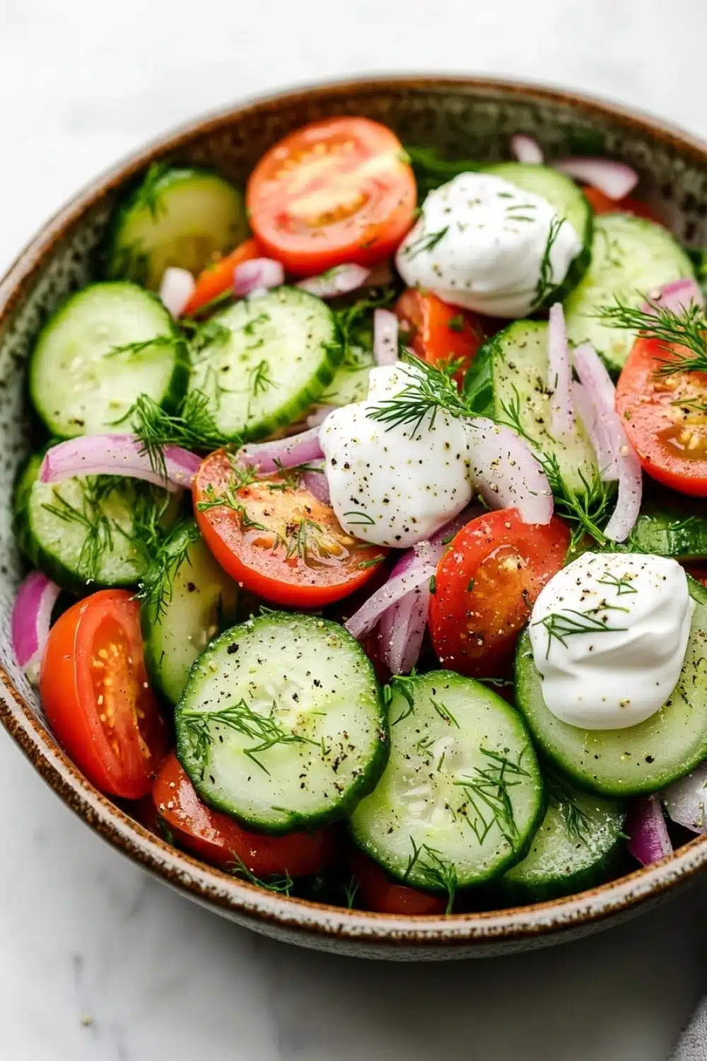 Cucumber tomato salad vinegar sour cream - the image is a close-up of a colorful salad in a bowl. the salad is made up of sliced cucumbers, red onions, cherry tomatoes, and dill leaves. the cucumbers are arranged in a circular pattern, with some overlapping each other. the tomatoes are bright red and appear to be ripe, while the onions are sliced into thin strips. the dill is scattered throughout the salad, adding a pop of green color to the dish. the bowl is sitting on a white marble countertop.