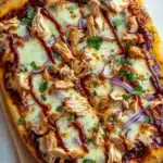 Detroit style bbq chicken pizza - the image is of a freshly baked pizza on a wooden cutting board. the pizza has a golden brown crust and is topped with melted cheese, shredded chicken, red onions, and a drizzle of bbq sauce. there are also a few sprigs of cilantro scattered on top of the pizza. the background is a white wooden table.