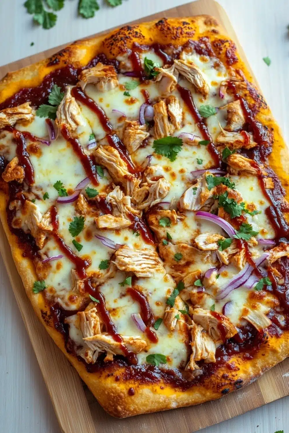 Detroit style bbq chicken pizza - the image is of a freshly baked pizza on a wooden cutting board. the pizza has a golden brown crust and is topped with melted cheese, shredded chicken, red onions, and a drizzle of bbq sauce. there are also a few sprigs of cilantro scattered on top of the pizza. the background is a white wooden table.
