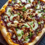 Detroit style bbq chicken pizza - the image is of a freshly baked pizza on a wooden cutting board. the pizza has a golden brown crust and is topped with melted cheese, shredded chicken, and chopped green onions. the cheese is melted and bubbly, and the chicken appears to be cooked until it is golden brown. the green onions are scattered on top of the cheese, adding a pop of color to the dish. the cutting board is placed on a dark wooden table, and there are a few sprigs of fresh green onions in the background.