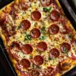 Detroit style pizza in cast iron - the image is of a freshly baked pepperoni pizza in a black cast iron skillet. the pizza is rectangular in shape and has a golden brown crust. it is topped with melted cheese, red pepperoni slices, and small chunks of tomato sauce. there are also some fresh basil leaves scattered on top of the pizza. the skillet is sitting on a dark countertop with a beige cloth napkin on the side.