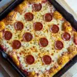 Detroit style pizza in cast iron - the image shows a rectangular pizza in a black cast iron skillet on a wooden cutting board. the pizza has a golden brown crust and is topped with melted cheese and pepperoni. the pepperoni is arranged in a circular pattern on top of the cheese. the skillet is sitting on a white countertop.