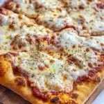 Detroit style pizza with frozen bread dough - the image shows a square pizza on a wooden cutting board. the pizza has a golden brown crust and is topped with melted white cheese and red sauce. the cheese appears to be melted and bubbly, and there are small black pepper sprinkled on top. the cutting board is placed on a white marble countertop.