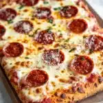 Detroit style pizza with frozen bread dough - the image is of a freshly baked pepperoni pizza on a wooden cutting board. the pizza is rectangular in shape and has a golden brown crust. the top of the pizza is covered in a generous layer of melted cheese, which is spread evenly over the crust. there are several slices of pepperoni arranged in a circular pattern, with some overlapping each other. the pepperoni is red and appears to be seasoned with herbs and spices. the cutting board is placed on a white marble countertop.