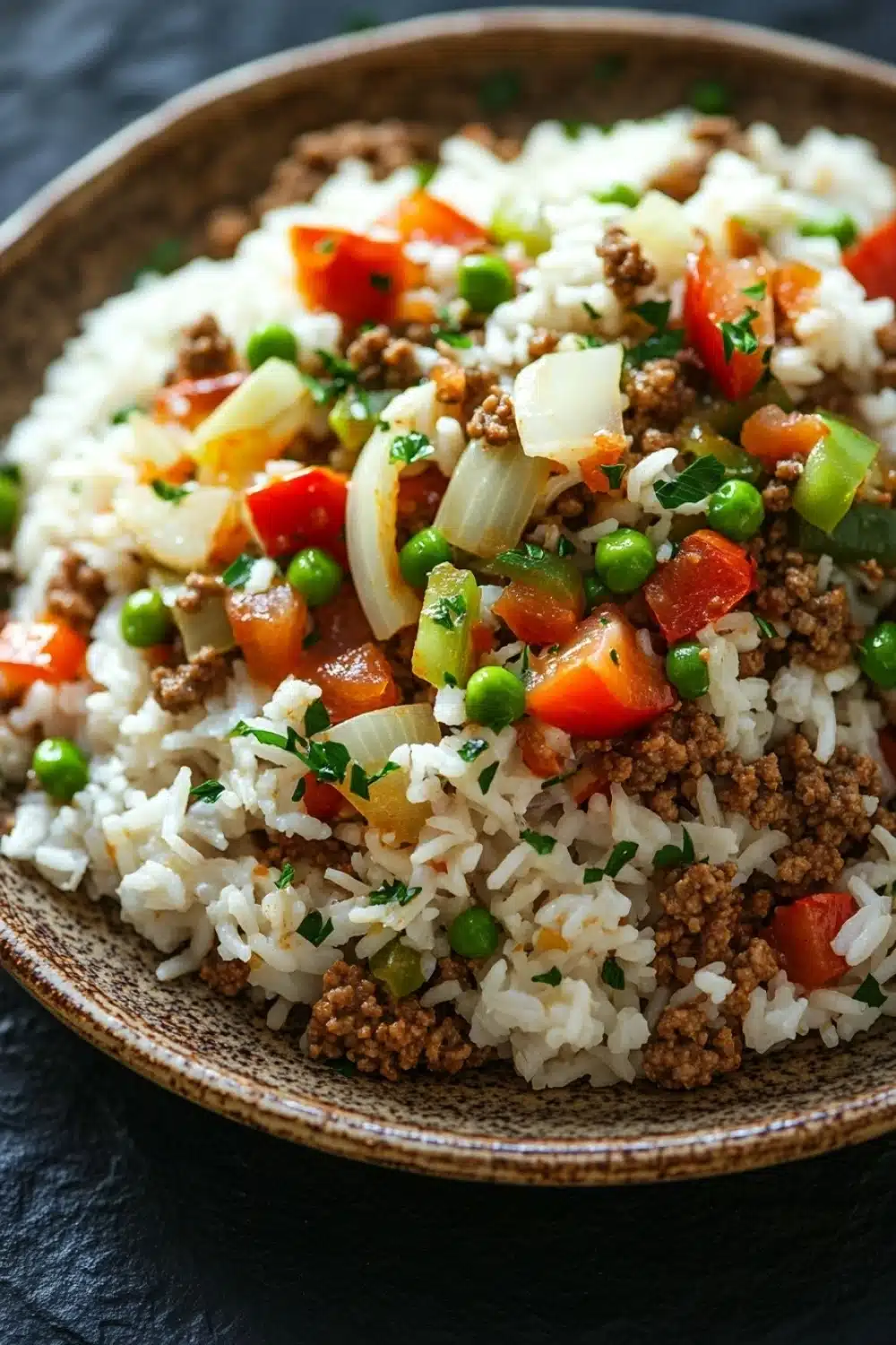 Dinner with ground beef and rice - the image is a close-up of a dish in a brown bowl. the dish appears to be a type of rice dish with ground beef, diced tomatoes, green peas, and onions mixed in. the rice is white and fluffy, and the ground beef is a dark brown color. the vegetables are bright red and appear to be diced into small cubes. the onions are cut into thin strips and are scattered throughout the dish. the bowl is sitting on a dark, textured surface, possibly a table or countertop.