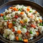 Dinner with ground beef and rice - the image shows a plate of fried rice with minced meat and vegetables. the rice is white and fluffy, and the meat appears to be ground beef. the vegetables include carrots, peas, and onions. the plate is brown and has a textured surface. the background is a dark wooden table.