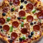 Diy sheet pan pizza - the image is of a pepperoni pizza on a wooden cutting board. the pizza is round and has a golden brown crust. it is topped with melted cheese, pepperoni slices, green bell peppers, and black olives. the pepperoni is arranged in a circular pattern on top of the cheese. the olives are scattered around the pizza, adding a pop of color to the dish. the cutting board is placed on a white wooden table.